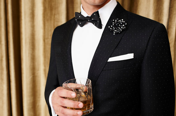 A man wearing a black tuxedo holding a glass of whisky.